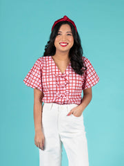 Woman wearing a red and pink checkered Nell blouse and white pants against a blue background