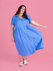 Woman wearing the Tilly and the Buttons Nell sewing pattern - a blue midi dress with short sleeves and a buttons down front, on a pink background.