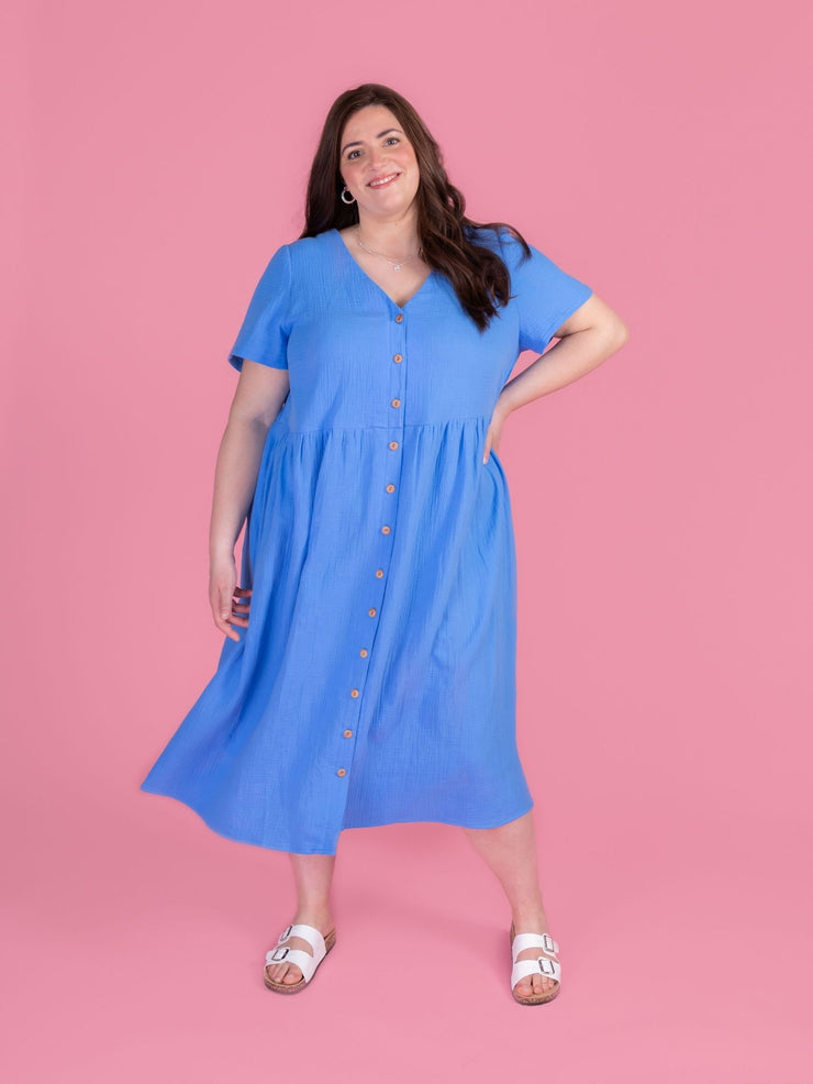 Woman wearing the Tilly and the Buttons Nell sewing pattern - a blue midi dress with short sleeves and a buttons down front, on a pink background.