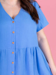Close up of woman wearing the Tilly and the Buttons Nell sewing pattern - a blue midi dress with short sleeves and a buttons down front, on a pink background.