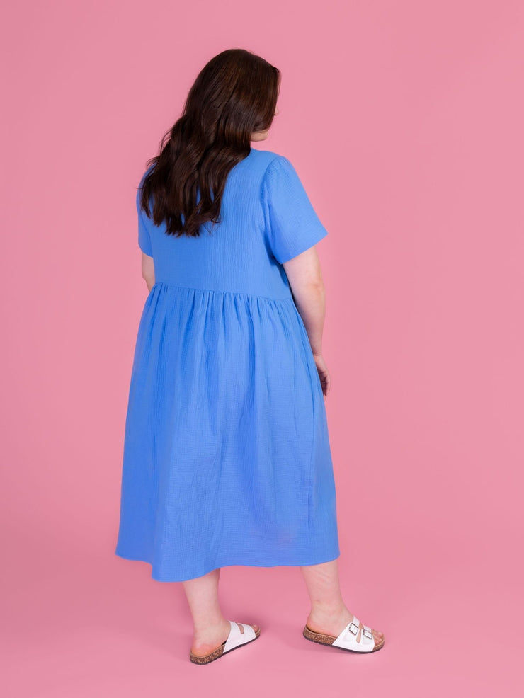 Woman wearing the Tilly and the Buttons Nell sewing pattern - a blue midi dress with short sleeves and a buttons down front, on a pink background.