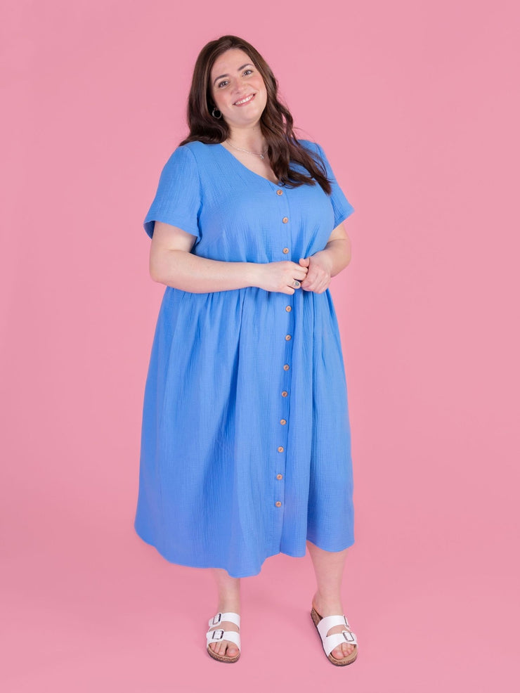 Woman wearing the Tilly and the Buttons Nell sewing pattern - a blue midi dress with short sleeves and a buttons down front, on a pink background.