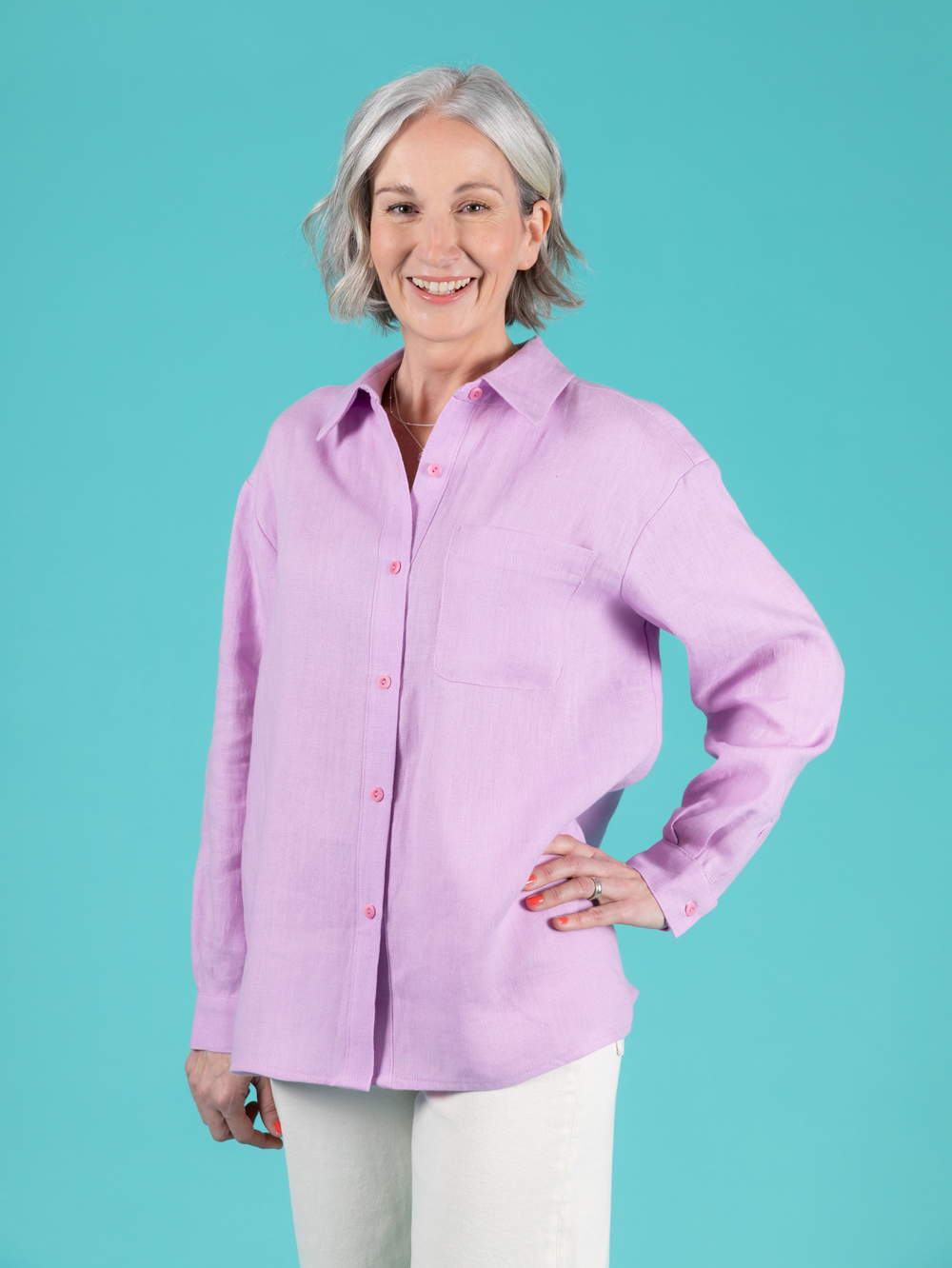 Woman wearing a lilac long sleeve Frida shirt and white jeans against a teal background.