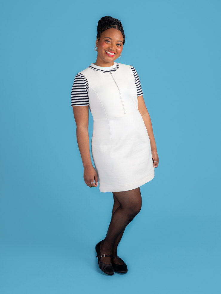 Woman wearing a white denim Noa pinafore dress with a black and white striped t shirt against a blue background.