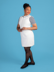 Woman wearing a white denim Noa pinafore dress with a black and white striped t shirt against a blue background.