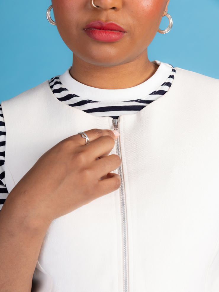 Close up of a woman wearing a white denim Noa pinafore dress with a black and white striped t shirt against a blue background.