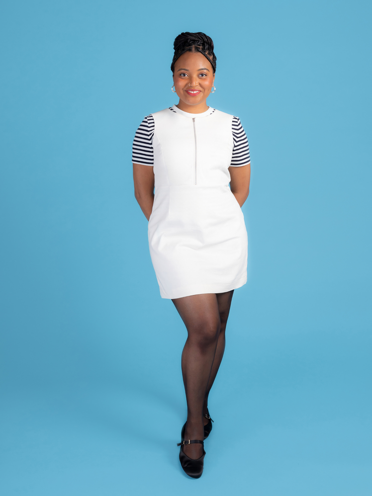 Woman wearing a white denim Noa pinafore dress with a black and white striped t shirt against a blue background.