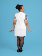 Back view of a woman wearing a white denim Noa pinafore dress with a black and white striped t shirt against a blue background.