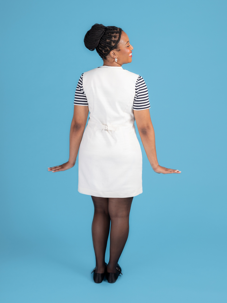 Back view of a woman wearing a white denim Noa pinafore dress with a black and white striped t shirt against a blue background.