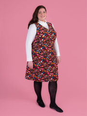 Woman wearing a multicoloured floral needlecord Noa pinafore dress with a long sleeved white roll neck top against a pink background.