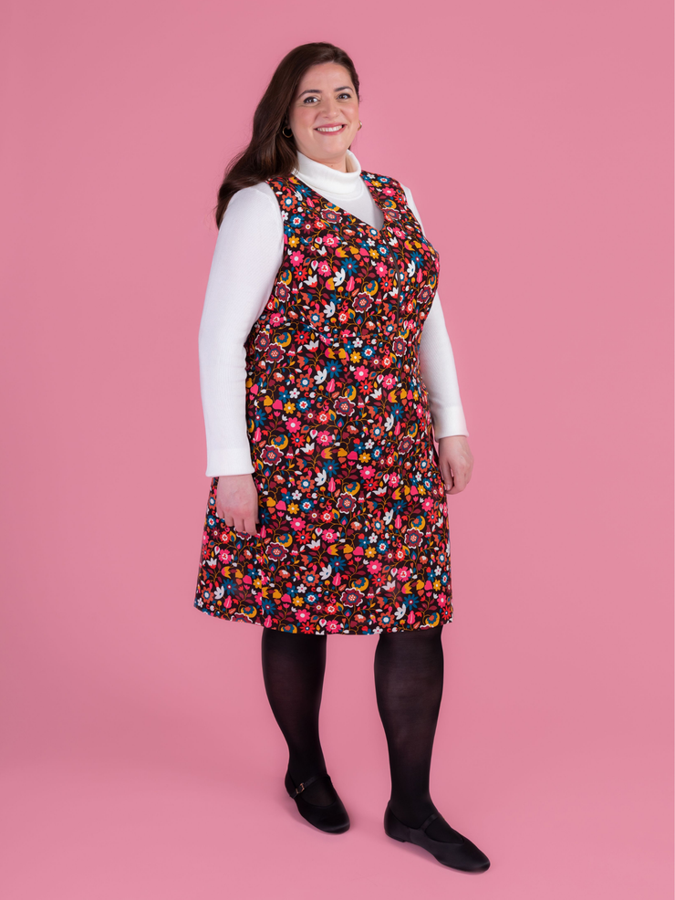 Woman wearing a multicoloured floral needlecord Noa pinafore dress with a long sleeved white roll neck top against a pink background.