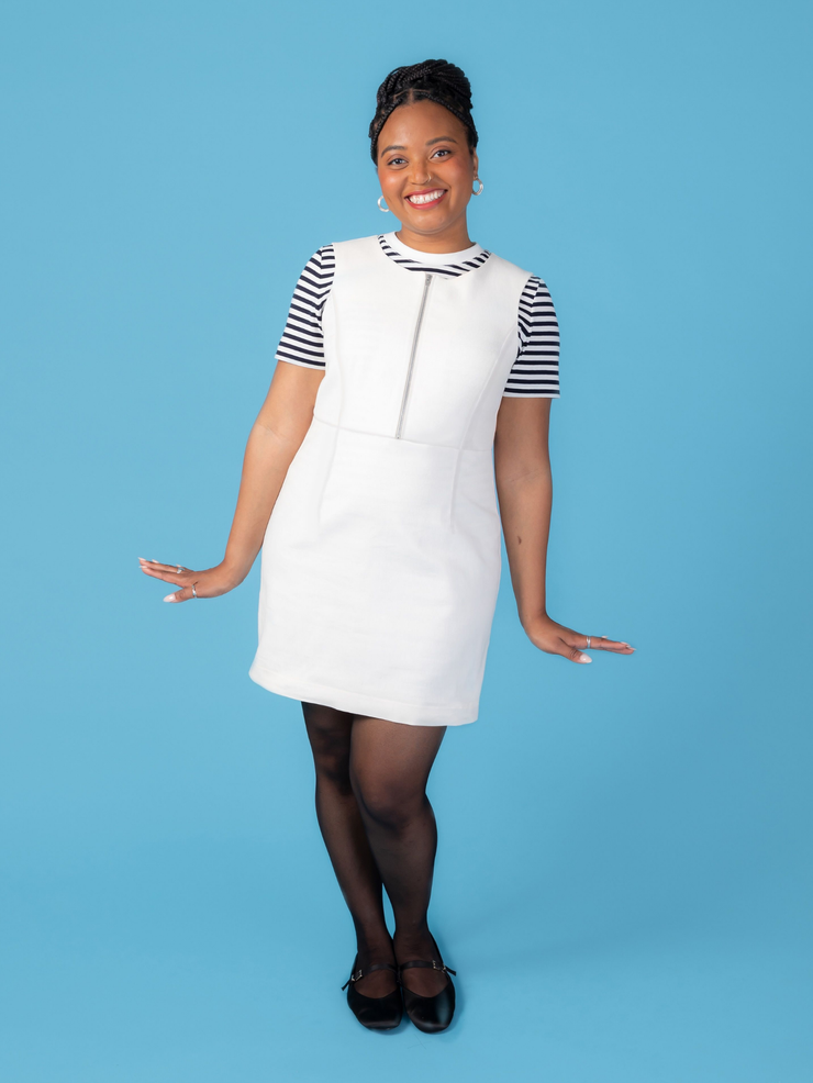 Woman wearing a white denim Noa pinafore dress with a black and white striped t shirt against a blue background.