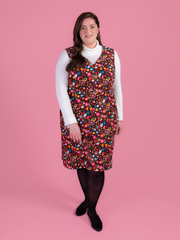 Woman wearing a multicoloured floral needlecord Noa pinafore dress with a long sleeved white roll neck top against a pink background.