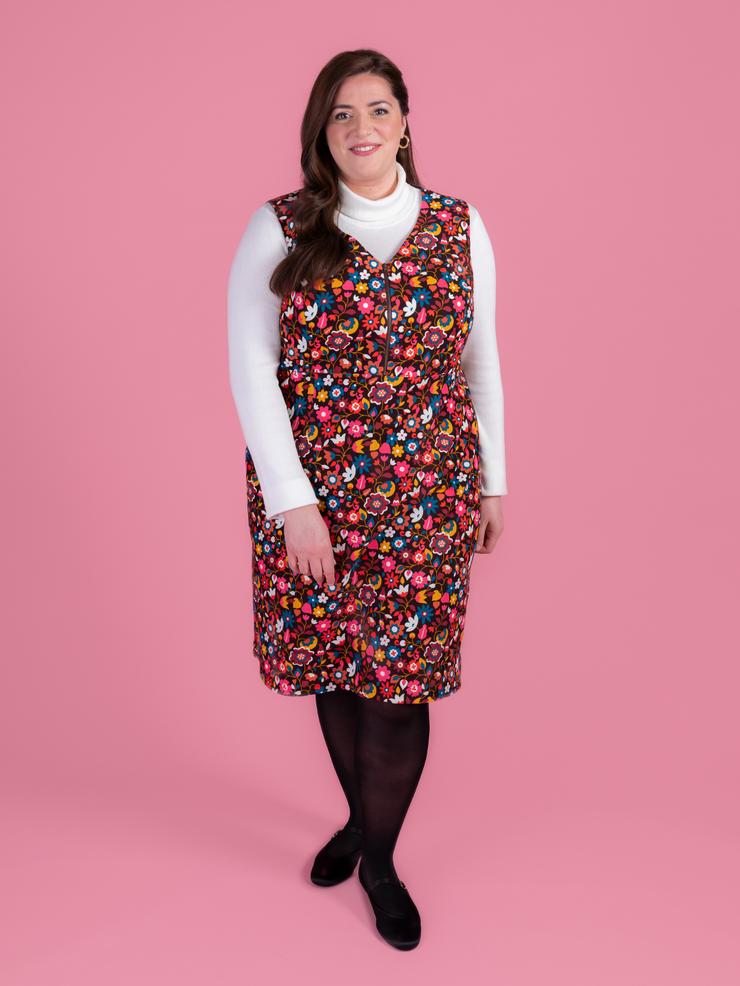 Woman wearing a multicoloured floral needlecord Noa pinafore dress with a long sleeved white roll neck top against a pink background.