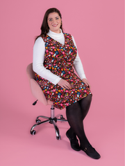 Woman sitting in a chair wearing a multicoloured floral needlecord Noa pinafore dress with a long sleeved white roll neck top against a pink background.