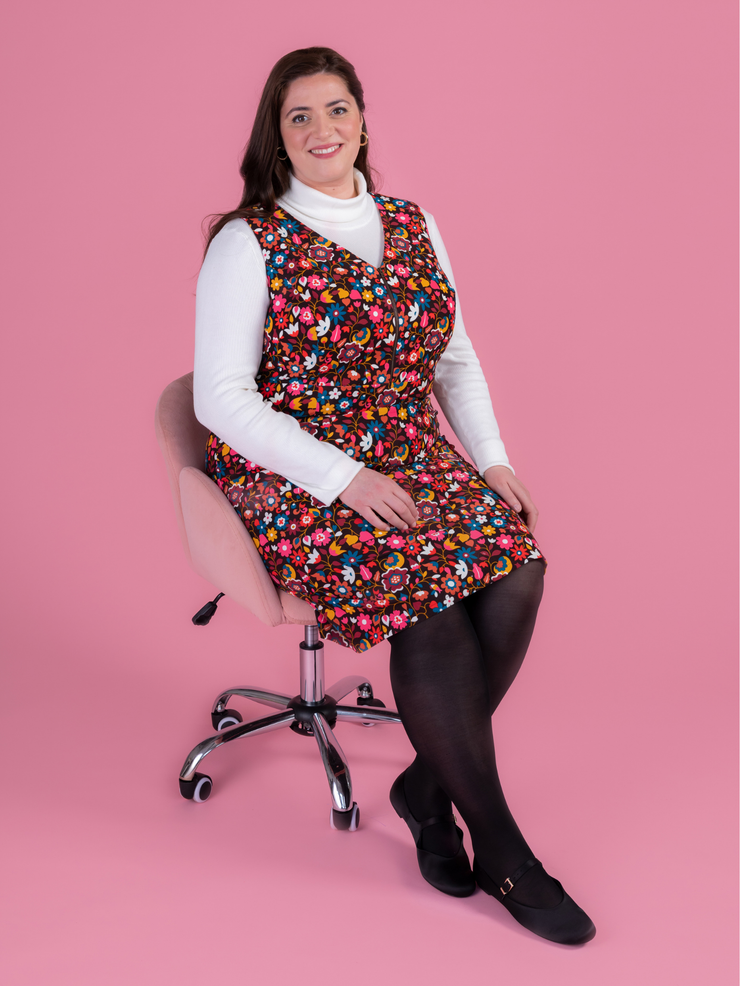 Woman sitting in a chair wearing a multicoloured floral needlecord Noa pinafore dress with a long sleeved white roll neck top against a pink background.