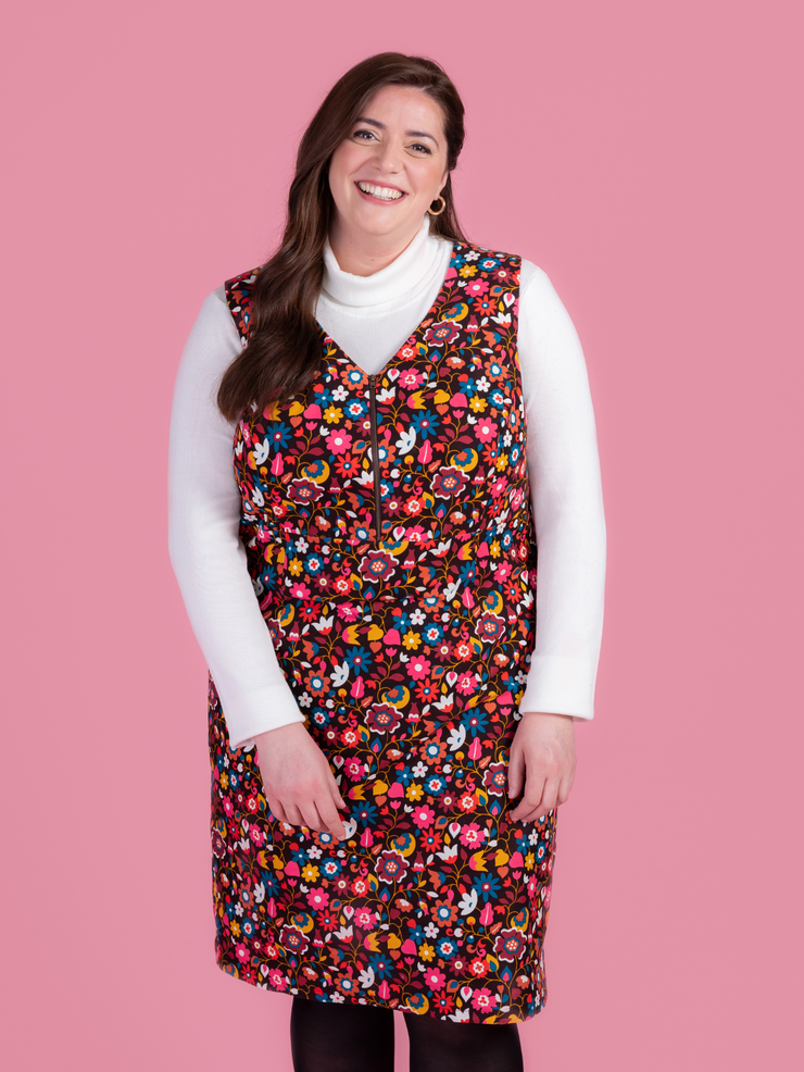 Woman wearing a multicoloured floral needlecord Noa pinafore dress with a long sleeved white roll neck top against a pink background.