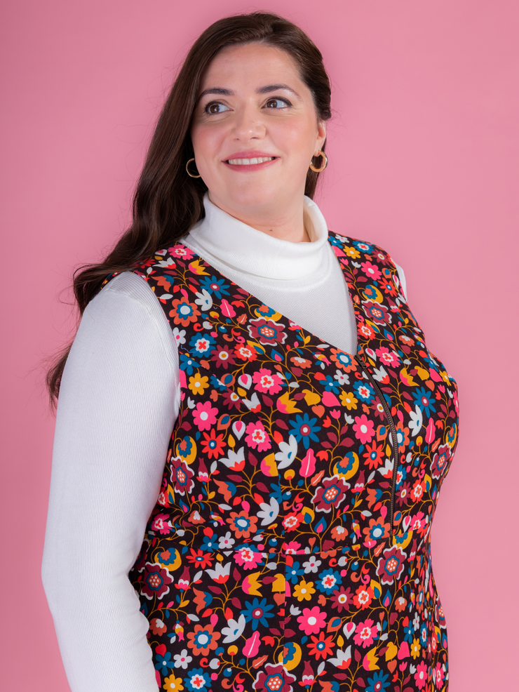 Woman wearing a multicoloured floral needlecord Noa pinafore dress with a long sleeved white roll neck top against a pink background.