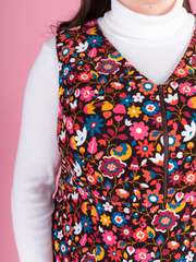 Close up of a woman wearing a multicoloured floral needlecord Noa pinafore dress with a long sleeved white roll neck top against a pink background.