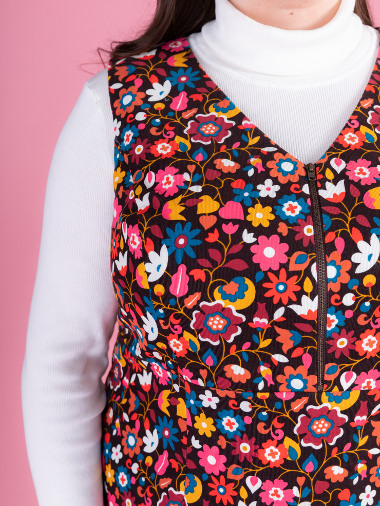 Close up of a woman wearing a multicoloured floral needlecord Noa pinafore dress with a long sleeved white roll neck top against a pink background.
