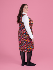 Woman wearing a multicoloured floral needlecord Noa pinafore dress with a long sleeved white roll neck top against a pink background.