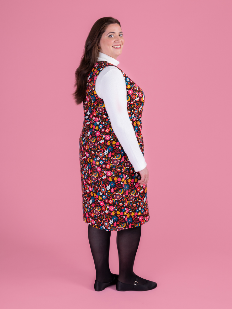 Woman wearing a multicoloured floral needlecord Noa pinafore dress with a long sleeved white roll neck top against a pink background.