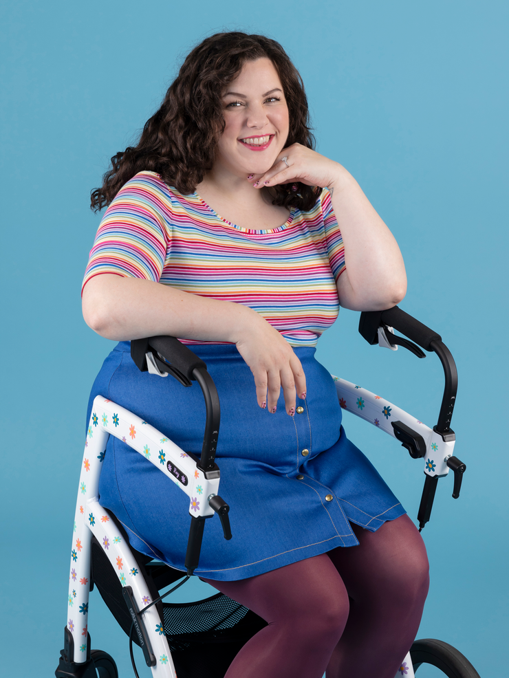 Model with curly brunette hair wears short sleeve rainbow stripe Agnes top while using mobility aid walker